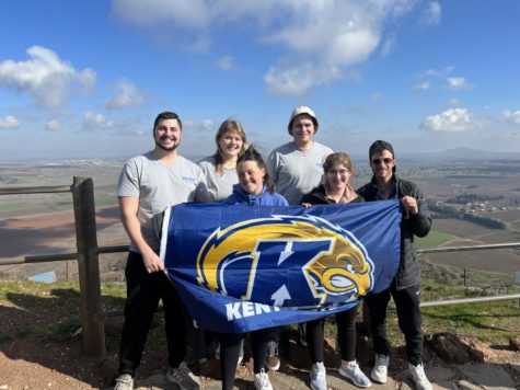 Six people stand behind a blue and gold Kent State flag they are holding. They are in Israel, on a hill with a valley behind them and blue skies above.