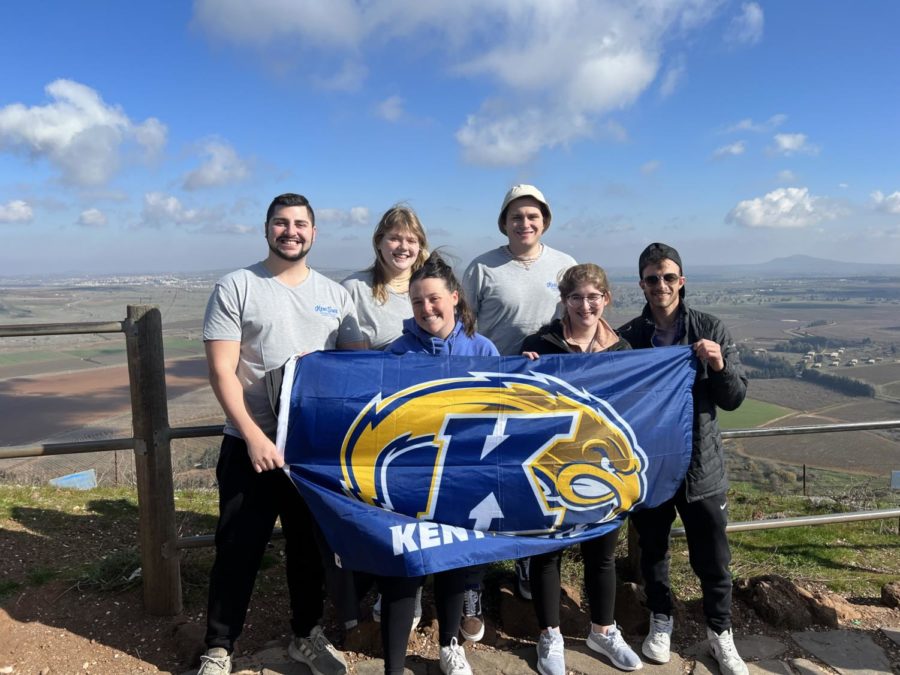 Six+people+stand+behind+a+blue+and+gold+Kent+State+flag+they+are+holding.+They+are+in+Israel%2C+on+a+hill+with+a+valley+behind+them+and+blue+skies+above.