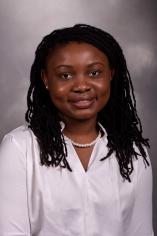 A Black woman with black hair is wearing a white shirt and smiling at the camera. She is in front of a gray backdrop.