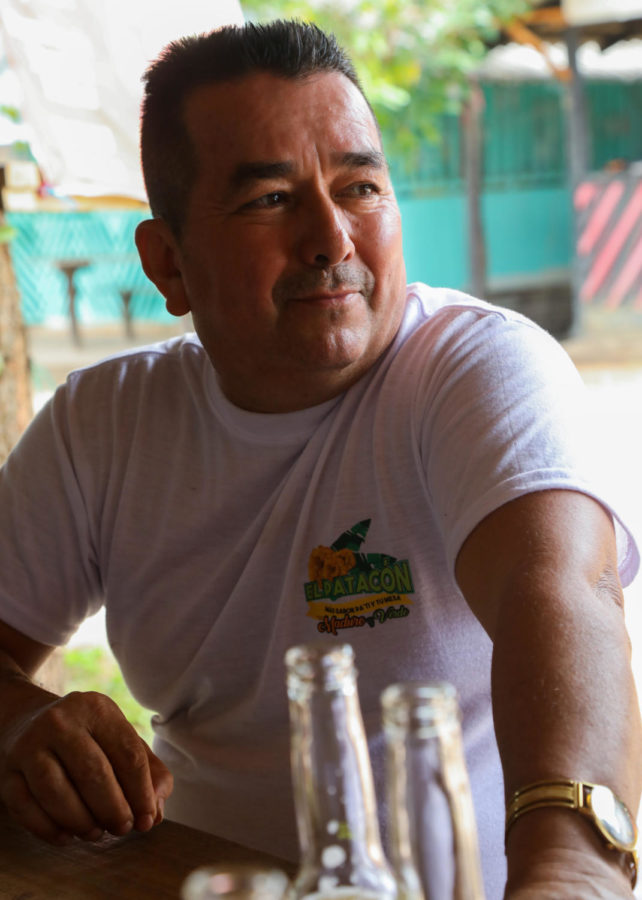 A Colombian man in a white shirt looks off to the right of the camera, leaning against a bar outside.