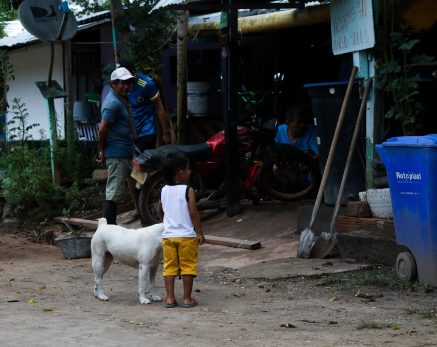 A child and goat stand in a dirt road outside a shop where two men work on a motorcycle.