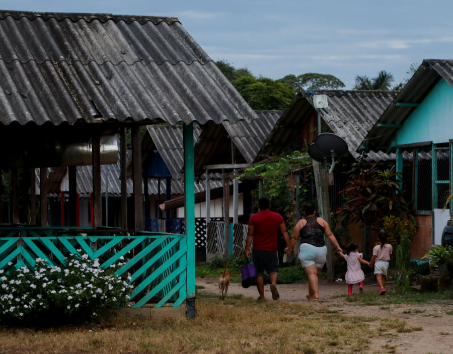Two adults walk with two children through the community of teal houses with thatched roofs.