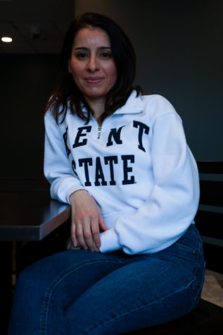 A young woman sits in a chair, leaning on the arm. She has dark hair and is wearing a white sweatshirt that says "Kent State" with dark wash jeans.